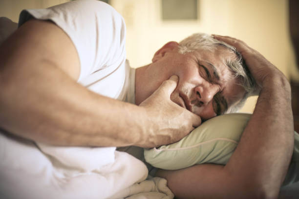 A man lies awake in bed, holding his jaw in discomfort due to tooth pain